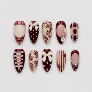 Può includere: Un set di dieci unghie finte a mandorla con una palette di colori bordeaux e crema. I disegni includono motivi in pizzo, pois bianchi, fiocchi color crema, volute dorate, piccoli bottoni, motivi a rombi, allacciature a corsetto e righe.