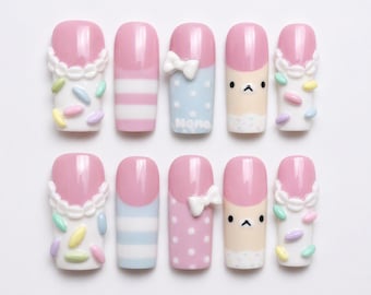 May include: A set of ten press-on nails featuring pink tips and various designs including white scalloped edges with pastel sprinkles, horizontal stripes, white polka dots with a 3D bow, and a cartoon bear face with the text Nomo on one nail.