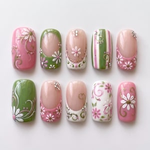 May include: A set of ten press-on nails featuring a variety of designs including pink and green bases, white daisy floral patterns, gold swirl accents, vertical green stripes, and small clear rhinestones arranged in two rows of five on a white background.