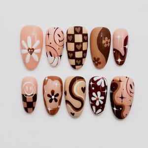 May include: A set of ten almond-shaped press-on nails featuring a brown, beige, and white color palette. Designs include checkered patterns with hearts, smiley faces, daisies, swirling abstract lines, and four-pointed star accents.