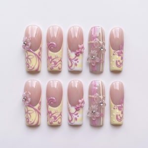 Könnte beinhalten: Ein Set aus zehn künstlichen Fingernägeln mit nudefarbener Basis, hellgelben Spitzen, violetten floralen Wirbelmustern und kleinen weißen Punkten. Einige Nägel sind mit dreidimensionalen violetten Blumenapplikationen und Strasssteinen verziert.