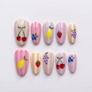 Op de afbeelding: Een set van tien kunstnagels met een glanzende afwerking en verticale pastelstrepen in roze, geel, blauw en groen. Elke nagel is versierd met een handgeschilderd fruitmotief, waaronder kersen, bosbessen, citroenen, aardbeien en druiven.