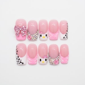 Peut inclure: Un ensemble de dix faux ongles avec une palette de couleurs rose et blanc. Les motifs incluent un nœud en strass rose, des cristaux transparents, des imprimés léopard, des rayures tigrées roses et un visage de chat avec un petit nœud rose.