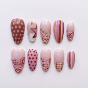 May include: A set of ten press-on nails featuring a variety of designs in shades of pink and brown. Patterns include polka dots, stars, leopard print, vertical stripes, swirls, geometric squares, and plaid, all set against a light pink base.