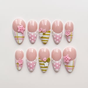 May include: A set of ten almond-shaped press-on nails featuring a nude base with white tips. Designs include pink and white plaid patterns, white polka dots on pink, and gold horizontal stripes. Embellishments include small pink star-shaped buttons.