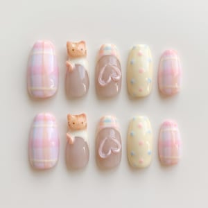 May include: A set of ten press-on nails featuring various designs including pink and blue plaid patterns, a three-dimensional orange and white cat figure, a raised heart shape on a translucent base, and a cream background with blue and pink dots.