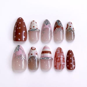 Puede incluir: Un juego de diez uñas postizas en forma de almendra con diversos diseños, incluyendo lunares blancos sobre fondo marrón, patrones geométricos en rosa y marrón, y detalles de volutas metálicas plateadas sobre una base translúcida brillante.