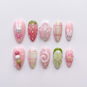 May include: A set of ten almond-shaped press-on nails featuring a pink base color. Designs include a white rabbit, a 3D strawberry, a pink flower, a pink and green plaid pattern, a dango skewer, a white swirl, a green flower, and a pink flower.