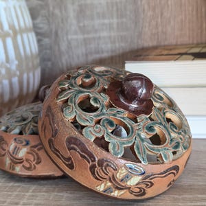 May include: A round, terracotta-colored ceramic lid featuring intricate, open-work scroll patterns highlighted in dark green and brown glaze. The lid has a smooth, dark brown circular knob at the center and decorative painted motifs along the base rim.