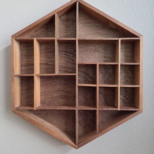 May include: A hexagonal wooden display shelf featuring a natural brown wood grain finish. The interior is divided into multiple compartments of varying sizes by thin wooden dividers, creating an asymmetrical grid pattern for organizing small items.