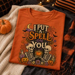 May include: An orange t-shirt featuring a graphic print with the text I PUT A SPELL ON YOU. The design includes two skeletons holding a glowing heart, surrounded by pumpkins, purple flowers, spiderwebs, bats, and a crescent moon.