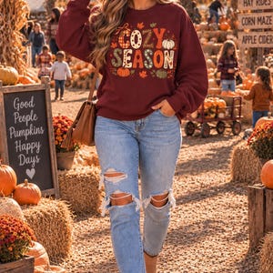 May include: A maroon long-sleeve sweatshirt featuring the text "COZY SEASON" in a decorative font, surrounded by illustrations of pumpkins and autumn leaves. The sweatshirt is paired with light blue denim jeans that have distressed knee detailing.