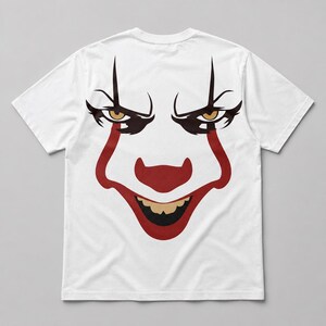 May include: A white short-sleeved t-shirt featuring a large graphic print of a clown face. The design includes sharp black eyebrows, yellow eyes, a prominent red nose, red mouth markings extending from the nose, and a wide grin with yellow teeth.
