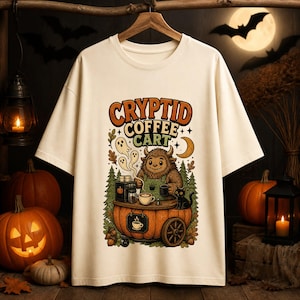May include: A cream-coloured t-shirt featuring a graphic of a furry creature operating a coffee cart made from a pumpkin. The text reads "CRYPTID COFFEE CART". The design includes small ghosts, a black cat, pine trees, and a crescent moon.