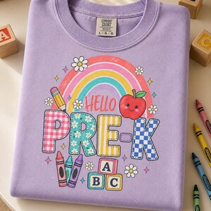 Könnte beinhalten: Ein hellviolettes T-Shirt mit einem Grafikdruck, der einen Regenbogen, einen lächelnden roten Apfel, einen Bleistift, Buntstifte und Alphabet-Blöcke zeigt. Der Text lautet HELLO PRE-K. Das T-Shirt hat einen Rundhalsausschnitt und ein Comfort Colors Etikett.