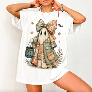 Könnte beinhalten: Ein weißes T-Shirt mit einer Grafik eines Geistes, der eine große Patchwork-Schleife und einen Patchwork-Umhang trägt. Der Geist hält einen dunkel-türkisfarbenen Eimer mit einem Halbmond- und Sternmotiv. Der Druck enthält kleine Sterne, eine Fledermaus.