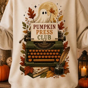 May include: A cream-colored crewneck sweatshirt featuring a graphic of a green vintage typewriter with orange pumpkin keys. A ghost holds a leaf above a paper reading PUMPKIN PRESS CLUB. The design includes autumn leaves, a book, and an inkwell.