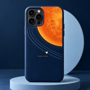 Könnte beinhalten: Eine dunkelblaue Smartphone-Hülle mit einer großen, texturierten orangefarbenen Planeten-Grafik in der oberen rechten Ecke. Dünne weiße Orbitallinien ziehen sich über die Oberfläche, ergänzt durch ein kleines Herz und den Text same orbit.