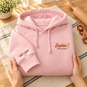 May include: A light pink hooded sweatshirt featuring the embroidered text Sophie BAKE STUDIO with a whisk icon on the chest and the text EST. 2021 embroidered on the sleeve cuff. The garment has a drawstring hood and long sleeves.