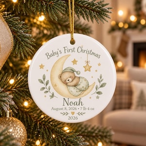 May include: A round white ceramic ornament hanging by a gold cord. The design features a sleeping teddy bear on a yellow crescent moon with stars, green leaves, and the text Baby's First Christmas, Noah, August 8, 2026, 7 lb 4 oz, and 2026.