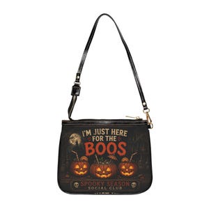 Könnte beinhalten: Eine schwarze Umhängetasche mit einem Grafikdruck von drei Kürbislaternen, die mit Getränken gefüllt sind. Der Text lautet I'M JUST HERE FOR THE BOOS und SPOOKY SEASON SOCIAL CLUB. Die Tasche hat einen schwarzen Riemen und einen Reißverschluss.