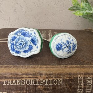 May include: Two ceramic drawer knobs with white surfaces and blue floral patterns. The larger knob has an irregular, scalloped edge with a green base, while the smaller knob is round with a scalloped border and a green base, both featuring blue painted flowers.