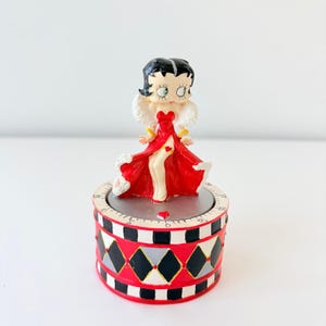 May include: A kitchen timer featuring a Betty Boop figurine wearing a red dress with white fur trim. The base is cylindrical with a black, white, and red diamond pattern and a silver top surface marked with black numbers for time settings.