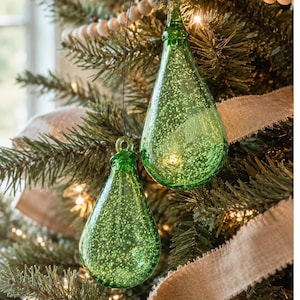 May include: Two teardrop-shaped glass ornaments in a vibrant green hue, featuring a textured surface filled with tiny air bubbles. Each ornament has a small glass loop at the top for hanging, suspended by a thin dark string against a pine tree.