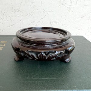 May include: A dark brown wooden display stand with a circular top and a carved, openwork base featuring floral-inspired patterns. The stand has a smooth, polished finish and rests on four small, curved feet, providing a stable platform for objects.