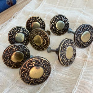 May include: A set of seven round cabinet knobs featuring a brass-toned center disc surrounded by a dark, antiqued metal frame with an intricate, repeating interlaced knot pattern. Each knob includes a metal mounting screw extending from the back.