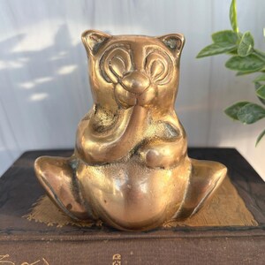 May include: A polished brass figurine of a seated bear with rounded ears and a smooth, reflective surface. The bear is depicted in a sitting position with its front paws raised toward its face, featuring a rounded belly and defined facial features.