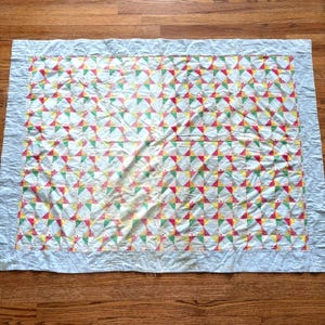 May include: A rectangular quilt featuring a repeating pinwheel pattern in shades of pink, green, and yellow on a white background. The central design is framed by a solid light blue border that extends to the edges of the fabric piece.