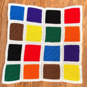 May include: A square crochet blanket featuring a grid of sixteen solid-colored squares in blue, purple, black, red, brown, yellow, green, and orange. Each square is separated by a thick, textured white border made of yarn stitches.
