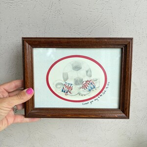 May include: A framed watercolor illustration featuring a soccer ball and a pair of athletic cleats inside a red oval border. The text "Commit your way to the Lord. Ps 37:5" is written in black ink on the lower right side of the off-white paper.