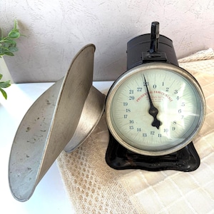 May include: A vintage black metal Prudential Family Scale with a round cream-colored dial featuring black numbers from 0 to 23 and a single black pointer. A detachable silver-toned metal scoop sits to the left of the scale base on a textured surface.