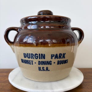 May include: A two-handled ceramic bean pot with a lid, featuring a two-tone brown and tan glaze. The front is imprinted with the dark blue text: DURGIN PARK MARKET • DINING • ROOMS U.S.A. The pot has a rounded body and a small knob on the lid.