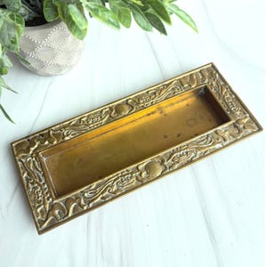 May include: A rectangular brass tray featuring an embossed border with intricate, repeating patterns of stylized creatures and floral motifs. The center of the tray is smooth and flat with a polished metallic finish, while the edges are raised.