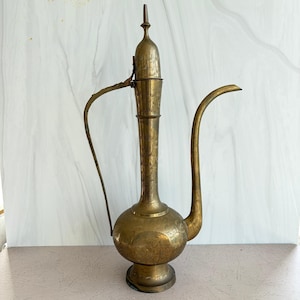 May include: A tall, antique-style brass ewer featuring an elongated neck, a slender curved spout, and a matching handle. The surface of the metal vessel is decorated with intricate, hand-etched floral patterns covering the body and neck.