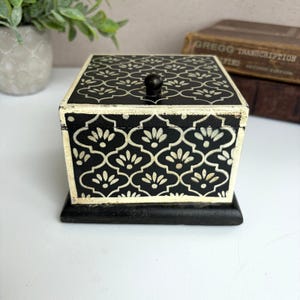 May include: A square wooden box with a black finish and a white Moroccan-style quatrefoil pattern. The box features a distressed, weathered edge effect, a solid black base, and a small, round black knob centered on the lid for opening the container.