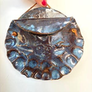 May include: A circular, dark blue glazed ceramic wall pocket featuring a hand-sculpted sun face design with rays extending to the scalloped edges. The piece includes a leather cord for hanging, with two small holes on the sides for the cord attachment.