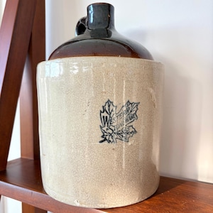 May include: A cylindrical stoneware jug with a cream-colored body featuring a fine crackle glaze and a dark brown glazed top. A blue maple leaf logo with the text Western Stoneware is printed on the front of the jug, which has a single handle.