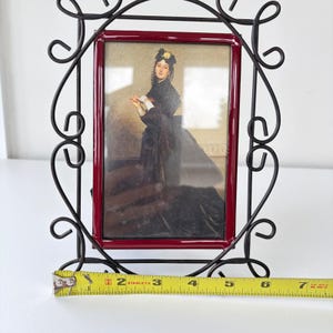 May include: A rectangular picture frame with a dark red inner border and a black metal scrollwork outer frame. A yellow measuring tape is positioned at the base, showing the frame width is approximately 7.5 inches. The frame holds a portrait print.
