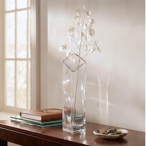May include: A tall, clear glass vase with a geometric, angular top edge. The vase has a square base and thick, transparent walls. A single, slender stem with multiple translucent, coin-shaped lunaria seed pods is placed inside the glass vessel.