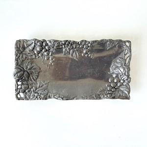 May include: A rectangular silver-toned metal tray featuring a raised, embossed border design of grape clusters and vine leaves. The central surface of the tray is smooth and reflective, while the edges are detailed with intricate botanical motifs.