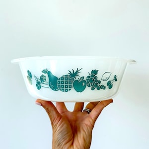 May include: A white glass mixing bowl featuring a repeating teal green fruit pattern around the exterior. The design includes illustrations of strawberries, a pear, a pineapple, an apple, grapes, a lemon, and cherries in a horizontal row.
