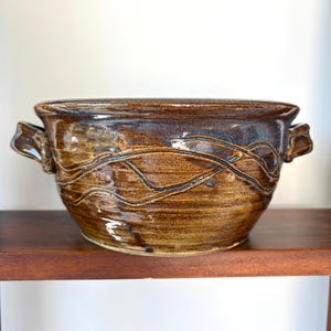 May include: A wide, round ceramic bowl with two side handles, featuring a glossy brown glaze with dark blue undertones and a raised, swirling line pattern that wraps around the exterior surface of the vessel, creating a textured appearance.