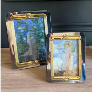 May include: Two rectangular picture frames with polished silver-tone outer borders and gold-tone inner borders featuring shell-like accents. The larger frame displays a water lily painting, while the smaller frame holds a painting of a person with a parasol.