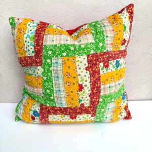 May include: A square throw pillow featuring a patchwork design composed of various fabric strips. The pattern includes bright yellow, green, and red floral, polka dot, and plaid prints stitched together in a grid-like arrangement.