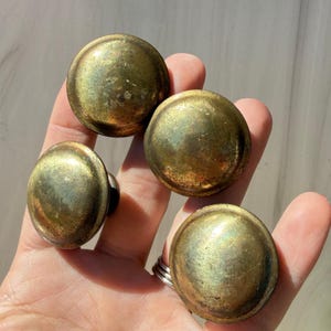May include: Four circular cabinet knobs with a weathered brass finish. Each knob features a smooth, domed surface with visible signs of patina and age, showing a metallic gold-toned texture across the rounded tops of the hardware pieces.