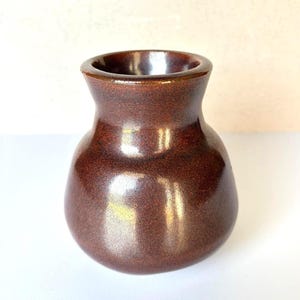 May include: A small, bulbous ceramic vase with a smooth, glossy finish in a deep, rich brown color. The vessel features a wide, rounded body that tapers into a narrower neck, ending in a circular opening with a thick, rounded rim.
